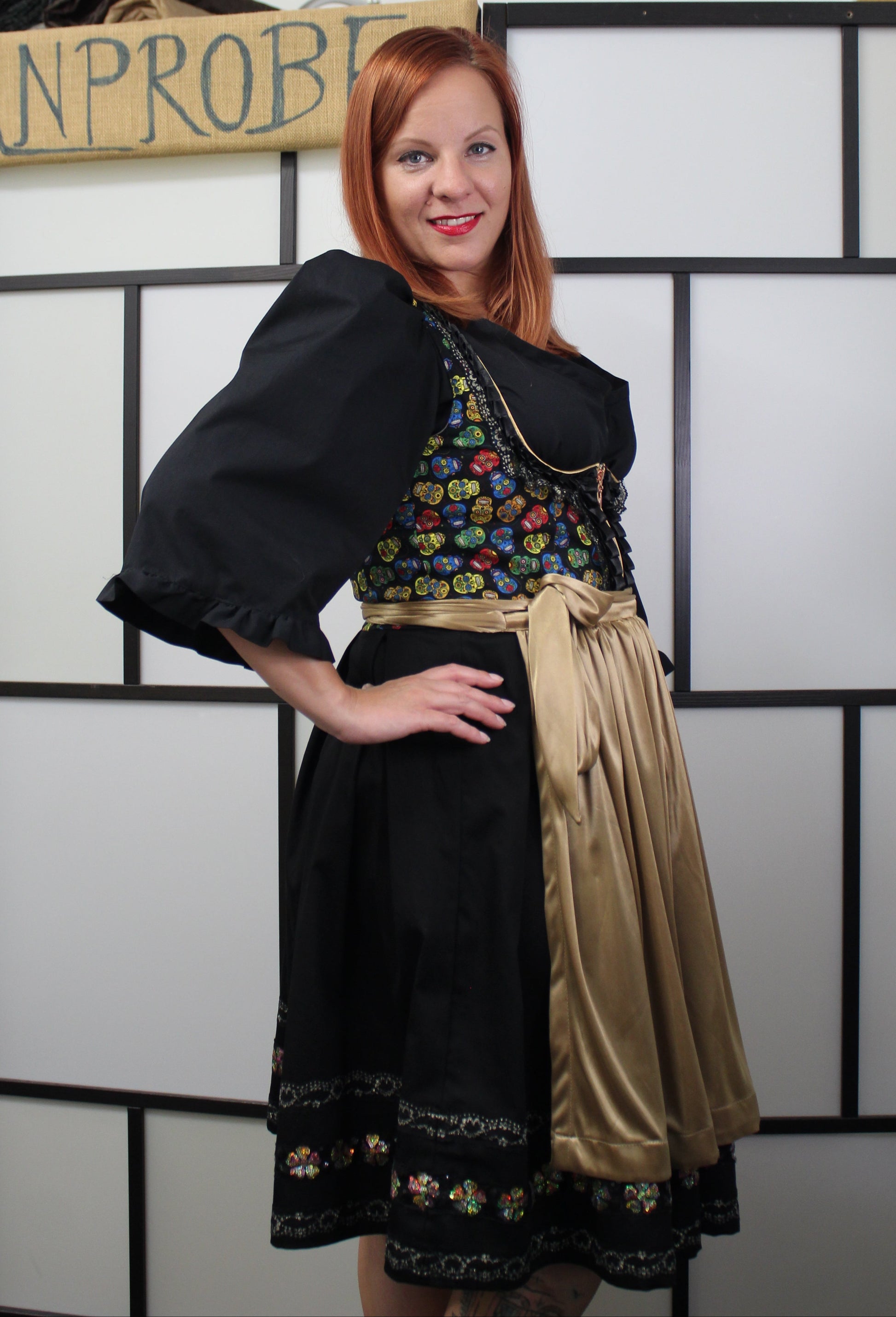 Model in a black and gold dirndl. Stoff mit klein Totenkopf-Muster und an Rock glänzende Blumen mit Spitzendetails.